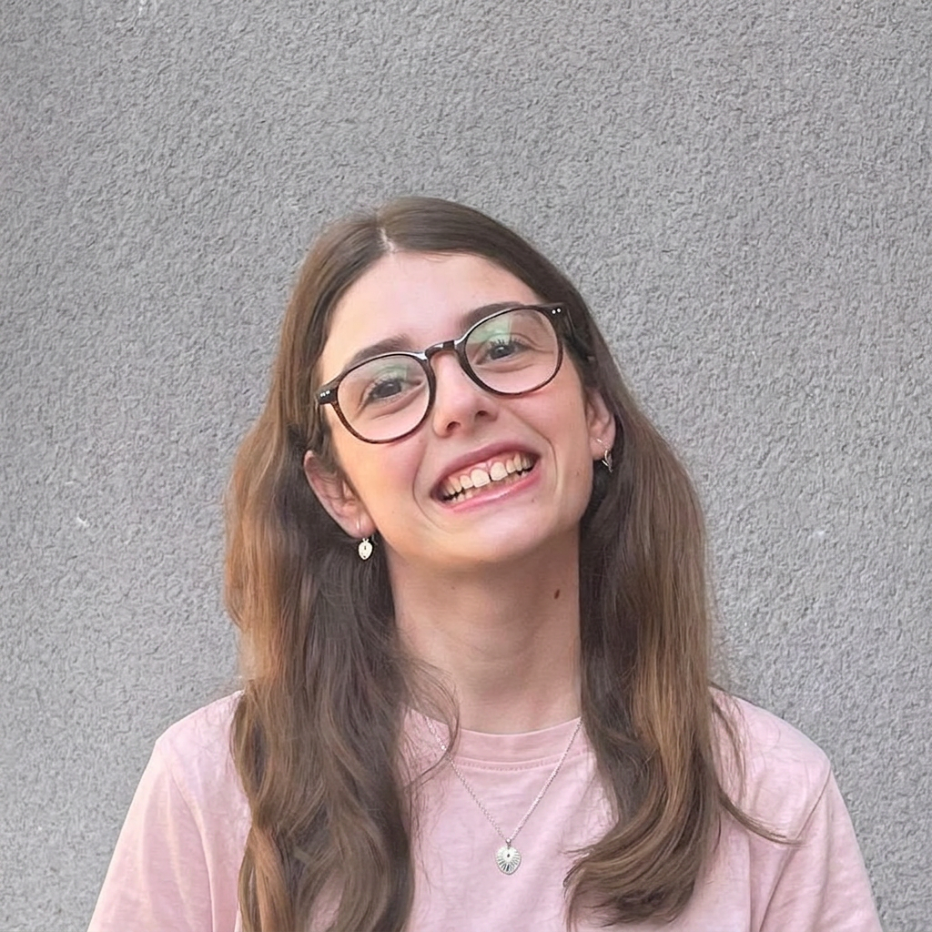 WhatsApp Image 2026-06-21 at 19.34.01 Smiling young woman with glasses and long hair, wearing a pink shirt, against a textured gray background.