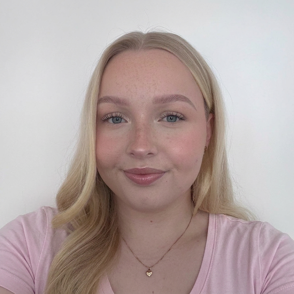 ANNA.nettisivu A young woman with long, wavy blonde hair and a subtle smile, wearing a pink shirt and a gold heart necklace, posed against a plain white background.