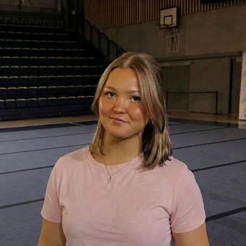 Näyttökuva 2026-06-21 194338 A young woman with shoulder-length hair wearing a pink t-shirt stands in a sports gymnasium with bleachers in the background.